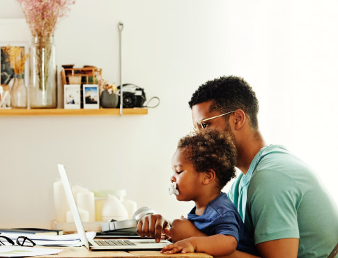 Father on the computer while holding a young child on his lap.